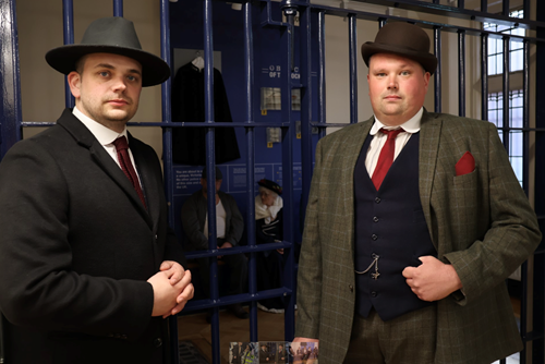 Two men in old-fashioned suits guard the entrance to a police cell