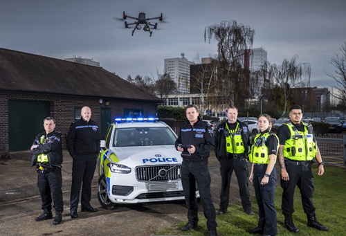 A team of police officers stand in front of a police car with a drone overhead