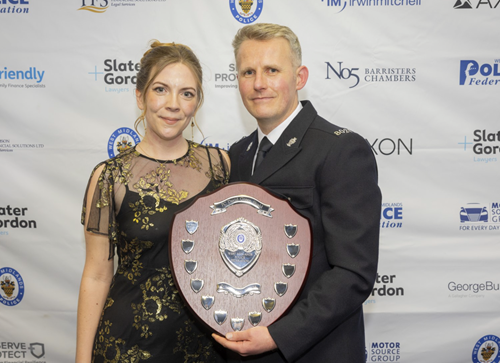 A police officer in black uniform holds a shield-dhaped award with his wife, who is wearing a black evening dress